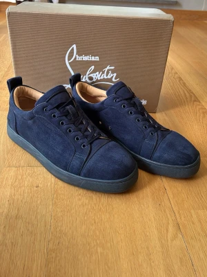 Mörkblåa sneakers i mocka - Snygga mörkblå sneakers i mocka från Christian Louboutin med svart sula och matchande mörkblå skosnören. Skorna har en stilren design med rund tå och diskreta sömmar. Insidan är ljusbeige och de har en låg profil som passar till många olika outfits.