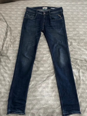 Mörkblå slim fit  jeans från Replay - Snygga mörkblå slim fit  jeans från Replay med klassisk femficksdesign och diskreta slitningar på låren. Jeansen har en smal passform och detaljerade bakfickor med Replay-logga. Perfekta för en trendig och avslappnad stil.