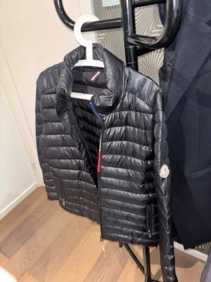 Svart dunväst från Moncler - Snygg svart dunväst från Moncler med quiltad design och hög krage. Västen har dragkedja framtill, två sidofickor och Moncler-logga på ärmen. Tillverkad i glansigt material med klassisk Moncler-detalj på insidan.