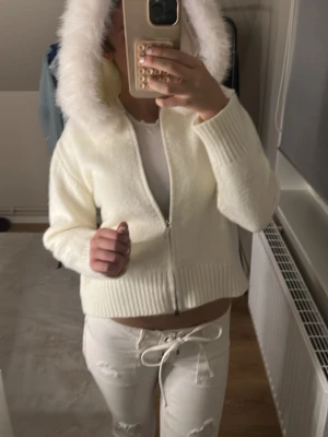 Vit hoodie med fuskpäls f - Mysig vit hoodie med dragkedja och stor luva kantad med fluffig fuskpäls. Tröjan är stickad med ribbade muddar vid ärmslut och nederkant. Perfekt för dig som gillar en clean och trendig look med lite extra cozy vibes.