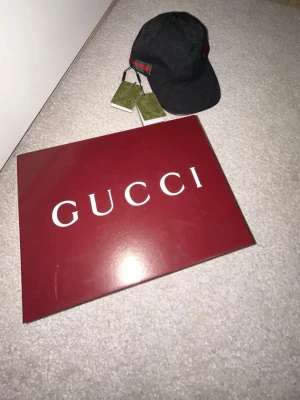 Svart keps från Gucci med logga - Snygg svart keps från Gucci med det klassiska GG-mönstret över hela kepsen. På sidan finns en grön och röd rand som ger en ikonisk touch. Kepsen är i tygmaterial och har justerbar passform. Perfekt för dig som vill ha en stilren accessoar. Fick den i maj la bara den i boxen och aldrig använde den nu säljer jag den har bara testats.