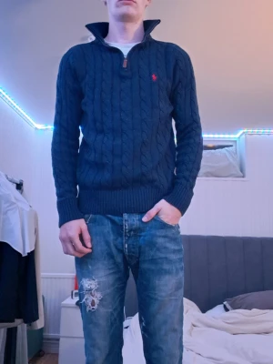 Mörkblå kabelstickad tröja Polo Ralph Lauren - Mörkblå kabelstickad tröja från Polo Ralph Lauren med halv dragkedja och hög krage. Tröjan har ett rött broderat logomärke på bröstet och ribbade muddar vid ärmslut och nederkant. Perfekt för dig som gillar klassisk och stilren look.