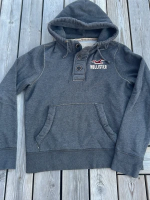 Grå hoodie från Hollister med knappar - Mysig grå hoodie från Hollister med huva och dragsko, tre knappar framtill och magficka. Broderad logga i vitt och vinrött på bröstet. Tjockt och mjukt material, perfekt för chill dagar. Snygga kontrastsömmar ger extra stil.