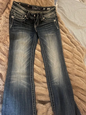 Blå bootcut jeans från Miss Me - Säljer ett par unika blå bootcut jeans från Miss Me. Använt 2 gånger och är som nya med tydliga slitningar och kontrastsömmar. Jeansen har fem fickor, dekorativa knappar och broderad logga på linningen. Perfekta för dig som gillar en avslappnad men snygg stil.