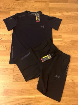 Svart träningsset från Under Armour - Snyggt svart träningsset från Under Armour med t-shirt och shorts. Båda plaggen har diskreta loggor och är tillverkade i ett lätt och snabbtorkande material, perfekt för löpning eller gym. T-shirten har rund hals och shortsen har resår i midjan.