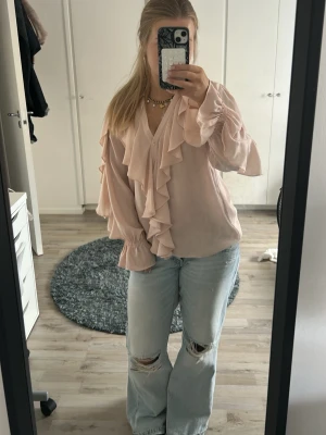 Ljusrosa volangblus med v-ringning - Supersöt ljusrosa blus med långa ärmar och volangdetaljer längs v-ringningen och framtill. Blusen har breda manschetter med resår och ett flowy, romantiskt fall. Perfekt att styla med jeans eller kjol för en trendig look.
