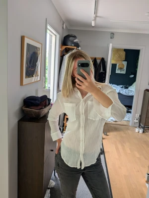 Vit skjorta - Säljer en vit skjorta i krinklat tyg med två bröstfickor och knappar framtill. Skjortan har lång ärm och klassisk krage, perfekt för en avslappnad men ändå stilren look. Materialet känns lätt och luftigt, vilket gör den skön att bära. Använd ett fåtal gånger 