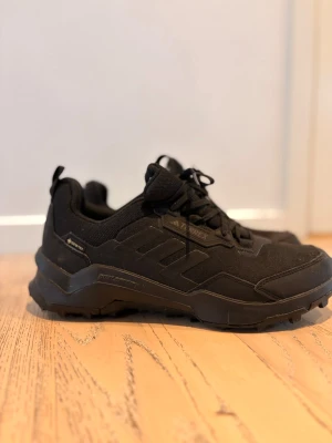 Svarta sneakers från Adidas - Säljer ett par helsvarta sneakers från Adidas med robust sula och diskreta detaljer. Skorna har en sportig design med mesh och syntetmaterial, samt snörning framtill. Perfekta för dig som gillar en clean och stilren look.