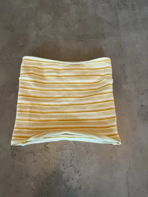 Randig bandeau tubtopp från Gina Tricot - Gul och vit randig bandeau tubtopp från Gina Tricot. Toppen är ribbad och har en mjuk, stretchig känsla som sitter snyggt på kroppen. Perfekt för varma dagar och enkel att matcha med jeans eller kjol.