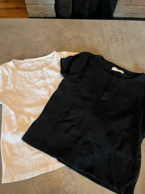 Svart och vit ribbad t-shirt med knappar - Två stilrena t-shirts i svart och vitt med ribbad struktur och knappar framtill. Båda har rund hals och korta ärmar, perfekta till jeans eller kjol. Materialet känns mjukt och stretchigt, troligen bomull eller bomullsblandning.