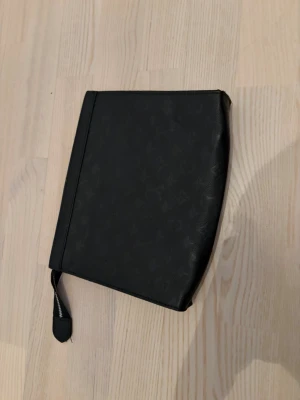 Svart clutch med monogrammönster - Snygg svart clutch med diskret monogrammönster och dragkedja. Väskan har en handledsrem och är tillverkad i ett slätt konstlädermaterial. Perfekt för att bära småsaker och accessoarer.