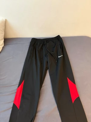 Svarta Nike träningsbyxor med röd detalj - Säljer ett par svarta träningsbyxor från Nike med röda detaljer på benen och vit logga på vänster sida. Byxorna har dragsko i midjan och smal passform, perfekta för gymmet eller löpning.