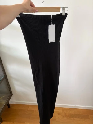Svart ribbad lång klänning från Bershka - Säljer en svart klänning från Bershka med ribbad struktur. Klänningen har en smal passform och är tillverkad i ett stretchigt material som sitter snyggt på kroppen. På baksidan av klänningen finns det en slits i mitten. Oanvänd och prislappen sitter kvar. 