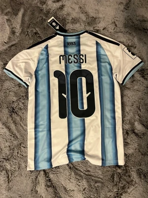 Argentina Messi matchtröja nr 10 - Säljer en officiell Argentina fotbollströja från Adidas med Messi och nummer 10 på ryggen. Tröjan har klassiska blå och vita ränder, svarta detaljer på axlarna och guldiga FIFA- och AFA-emblem på bröstet. Perfekt för dig som älskar fotboll och Messi. Passar M bra