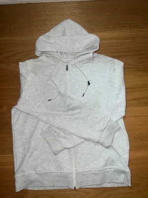 Ljusgrå zip hoodie från Polo Ralph Lauren - Snygg ljusgrå hoodie med dragkedja från Polo Ralph Lauren. Klassisk huva med dragsko, två fickor framtill och den ikoniska mörkblå loggan broderad på bröstet. Perfekt för en chill och stilren look. Mjuk bomullsblandning för extra komfort.