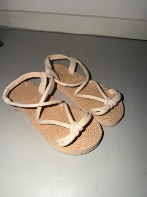 Beige sandaler med repdetaljer - Säljer ett par trendiga beige sandaler med flätad sula och repband som går över foten och runt ankeln. Sandalerna har öppen tå och en avslappnad design med knutdetaljer framtill. Perfekta för sommaren och ger en bohemisk känsla.