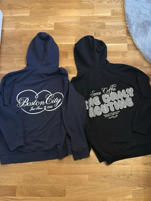 Två hoodies med tryck, blå & svart, lager 157 - Säljer två hoodies, en marinblå med 'Boston City Just Since 1998' tryck i vitt och en svart med texten 'Some Coffee The Daily Routine' på ryggen. Båda har huva med dragsko och ribbade muddar. Perfekta för en chill och avslappnad stil.
