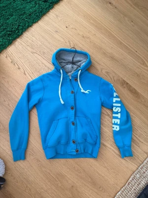 Blå Hollister hoodie med knappar - Snygg blå hoodie från Hollister med knappar framtill, vita dragsnören och grått foder i huvan. Stor logga på ena ärmen och klassisk Hollister-fågel på bröstet. Två fickor fram och ribbade muddar. Väldigt bra skick. Stolen Svara