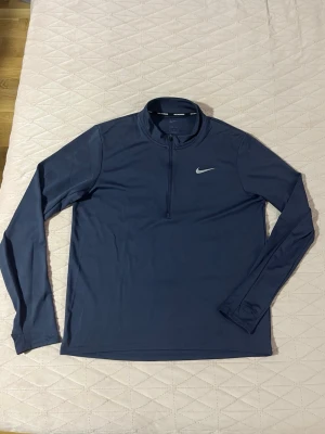 Mörkblå långärmad träningströja Nike - Snygg mörkblå långärmad träningströja från Nike med halv dragkedja framtill och diskret Nike-logga på bröstet. Tillverkad i ett mjukt och stretchigt funktionsmaterial som passar perfekt för träning eller löpning.