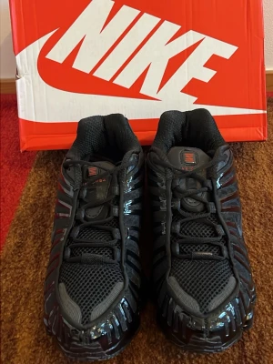 Nike Shox TL svarta sneakers - Nike Shox TL sneakers i helsvart med glänsande detaljer och meshpaneler. Skorna har den ikoniska Shox-sulan med fjädrande pelare och ett sportigt, futuristiskt utseende. Swoosh-logga på sidan och Shox-märke på plösen. Grymt snygga till streetwear!