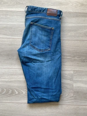Blå raka jeans med slitningar  - Säljer ett par klassiska blå jeans med rak passform och fem fickor. Jeansen har en snygg skinnpatch i midjan och slitningar framtill för en cool, avslappnad look. Perfekta till vardagsstilen och tillverkade i denim. Riktigt schyssta jeans från Lindberg ordinarie pris 1299. Med snygga slitningar 