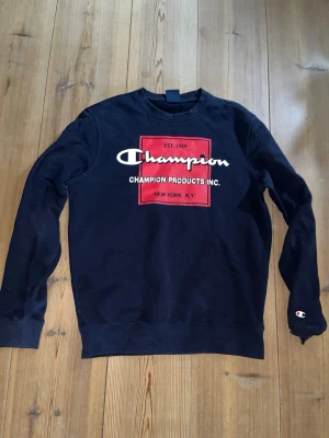 Marinblå sweatshirt från Champion - Cool marinblå sweatshirt från Champion med stort rött och vitt tryck på bröstet där det står 'Champion Products Inc.' och 'New York N.Y.'. Tröjan har rund halsringning och ribbade muddar vid ärmar och nederkant. Perfekt för en avslappnad streetwear-look.    Nypris:649kr.      Mitt pris:349kr