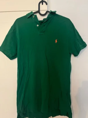 Grön piké från Polo Ralph Lauren - Säljer en grön pikétröja från Polo Ralph Lauren i slim fit. Klassisk krage, kortärmad och med den ikoniska broderade loggan på bröstet. Tillverkad i bomull och har knappar vid halsen. Perfekt för en clean och stilren look.