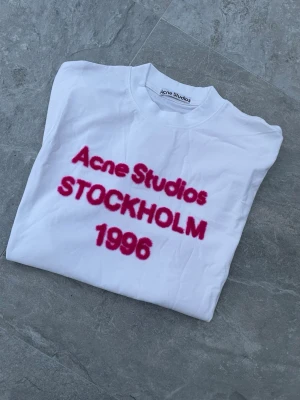 Acne Studios T-Shirt - Supersnygg vit Acne T shirt till tjej med fluffig rosa text. Storlek S, passformen är oversized så passar även större storlek. Perfekt att matcha till trendiga outfits. Skickar snabbt!