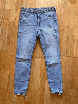 Ljusblå curvy jeggings från H&M - Säljer ett par ljusblå curvy jeggings från H&M med hög midja och ankellängd. Jeansen har slitningar framtill och råa, fransiga benslut för en trendig look. Tillverkade i stretchigt denim-material för extra komfort.