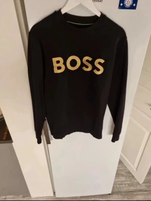 Svart sweatshirt från Hugo Boss - Säljer en svart sweatshirt från Hugo Boss med stor guldig text 'BOSS' på bröstet. Tröjan har rund halsringning. Tillverkad i mjukt material som passar perfekt till en avslappnad stil. Boss påse kommer självklart med på köpet skriv vid fundering