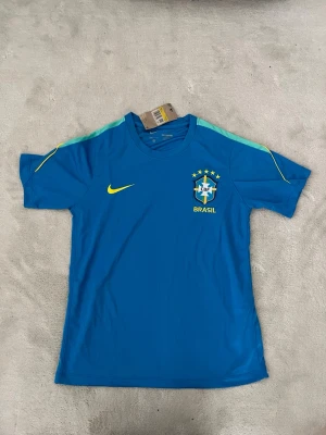 Blå Brasilien Nike träningströja - Snygg blå träningströja från Nike med Brasilien-logga och texten BRASA i gult på ryggen. Tröjan har korta ärmar, rund hals och ljusgröna detaljer på axlarna. Perfekt för fotbollsträning eller att representera ditt favoritlandslag.