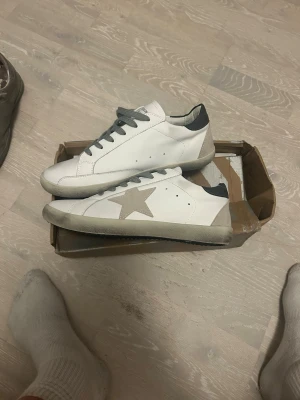 Vita golden goose - Snygga vita golden goose med grå stjärndetalj på sidan och grå skosnören. Skorna har en låg profil och grå hälflik. Perfekta för en avslappnad och trendig stil. 