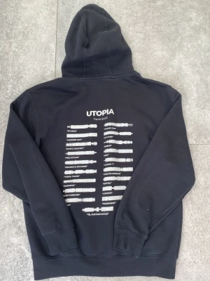 Svart Utopia hoodie Travis Scott - Säljer en svart hoodie med stor huva och magficka. På ryggen finns ett vitt tryck med texten 'UTOPIA Travis Scott' och en lista med låttitlarna. Perfekt för dig som gillar streetwear eller rap