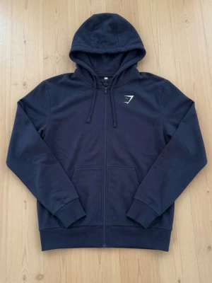 Mörkblå hoodie från Gymshark - Snygg mörkblå hoodie från Gymshark med dragkedja framtill och två fickor. Hoodien är i XS men passar även S och till och med M om du är lite kortare. Har justerbar huva med snören och Gymshark-logga på bröstet. 