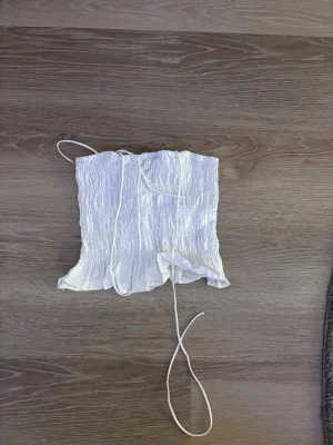 Vit croppad topp med snörning - Säljer en vit croppad topp med smock och långa snören framtill. Toppen har volangkant nedtill och är perfekt för sommaren. Xs, från H&M Helt ny och oanvänd.