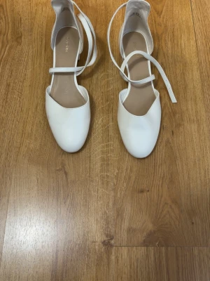 Vita ballerinaskor med remmar - Säljer ett par vita ballerinaskor med låga klackar och smala remmar som korsas över foten. Skorna har rundad tå och är tillverkade i ett slätt material som ser ut som konstläder. Perfekta för en stilren och trendig look.