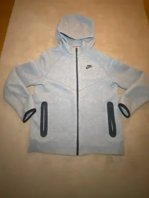 Grå Nike tech hoodie  - Snygg ljusgrå hoodie från Nike med hel dragkedja framtill och två svarta fickor med dragkedja. Tröjan har huva och svarta detaljer vid ärmsluten. Perfekt för en sportig och avslappnad stil.