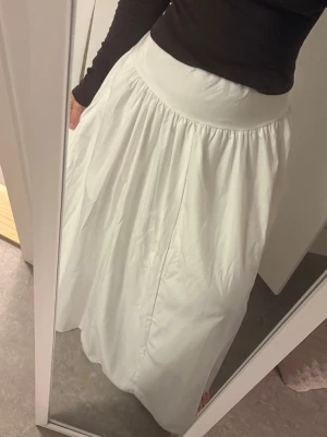 Vit lång kjol med hög midja - Säljer en lång, vid kjol i vitt med hög midja. Kjolen har en enkel och stilren design med lätt rynkad midja för extra volym. Perfekt för dig som gillar en flowy och elegant look.