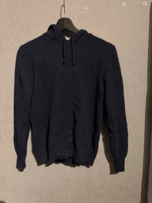Kashmir hoodie - En enkel och stilren mörkblå hoodie med huva och dragsko. Hoodien har långa ärmar och ribbade muddar vid ärmslut och nederkant. Perfekt för en avslappnad look och passar till det mesta. Materialet ser ut att vara mjukt och bekvämt.