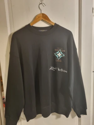 Svart Louis Vuitton sweatshirt - Cool svart sweatshirt från Louis Vuitton med stor turkos och vit grafisk logga på ryggen och mindre logga framtill. Tröjan har rund hals, ribbade muddar och avslappnad passform. Perfekt för dig som gillar streetwear och exklusiva märken. 