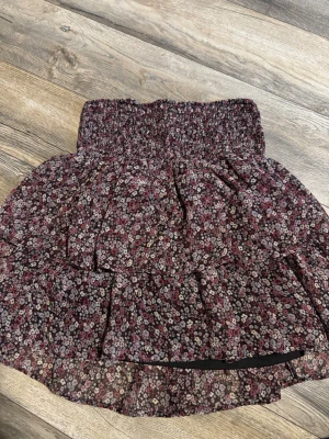 Blommig kortkjol från Kappahl - Supersöt kortkjol från Kappahl med blommigt mönster i lila, rosa och vitt på mörk botten. Kjolen har bred smock i midjan och volangdetaljer som ger en luftig och flowy känsla. Perfekt för dig som gillar en feminin och trendig stil.