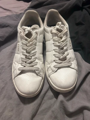 Vita låga sneakers från Björn Borg - Klassiska vita sneakers från Björn Borg med lågt skaft och snörning. Skorna har en stilren design med diskret logga på sidan och är tillverkade i slätt syntetmaterial. Perfekta för en clean och enkel look.