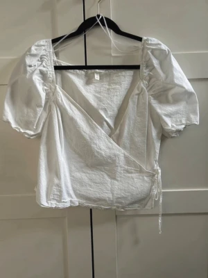 Topp - Söt vit omlottopp med puffiga korta ärmar och knyt i sidan. Toppen har en v-ringad skärning och är gjord i ett lätt bomullstyg som passar perfekt till varma dagar. Enkel men trendig design som är lätt att styla.