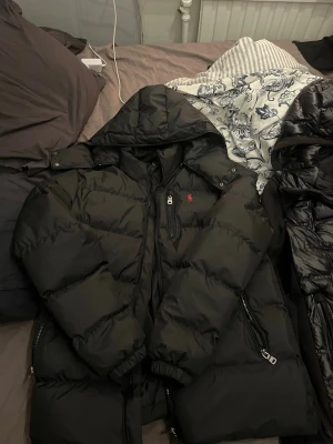 Ralph Lauren Jacka - Säljer en svart pufferjacka från Polo Ralph Lauren med huva och dragkedja framtill. Jackan har två sidofickor med dragkedja och den klassiska röda loggan på bröstet. Perfekt för kalla vinterdagar och riktigt skön att bära.
