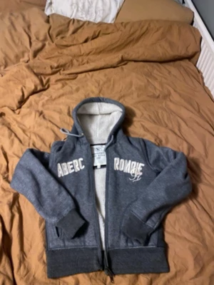 Grå hoodie med dragkedja och huva - Mysig grå hoodie med dragkedja och vit text på bröstet. Hoodien har ribbade muddar, fickor på sidorna och en fodrad insida i ljusare tyg. Perfekt för chill dagar och enkel att matcha med jeans eller mjukisbyxor. Står storlek L men krympt i tvätten så skulle säga storlek s