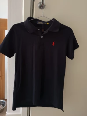 Marinblå pikétröja från Polo Ralph Lauren - Klassisk marinblå pikétröja från Polo Ralph Lauren med korta ärmar och röd broderad logga på bröstet. Tröjan har krage och knappar framtill, perfekt för en stilren och avslappnad look.