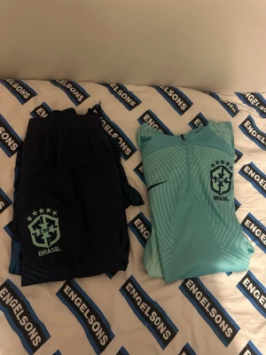 Nike Brasil Dress - Snyggt träningsset från Nike med Brasil-logga. Setet består av ett par mörkblå shorts och en ljusblå långärmad träningströja med halv dragkedja och grafiskt mönster. Perfekt för fotbollsträning eller gymmet. Materialet är lätt och andas.