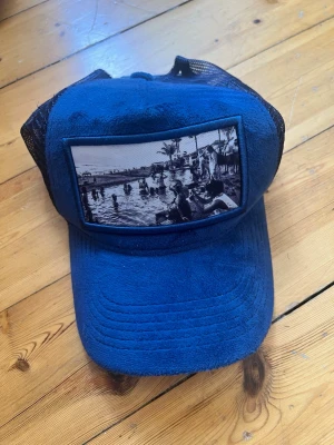 Blå truckerkeps med strandmotiv - Snygg blå truckerkeps med svart mesh baktill och ett stort tygmärke framtill med svartvitt strandmotiv. Kepsen har böjd skärm och justerbar snapback. Perfekt för dig som vill sticka ut med en unik accessoar.