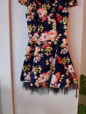 H&R Klänning Small - Snygg marinblå kortklänning med färgglatt blommönster i rosa, vitt och grönt. Klänningen har korta ärmar, v-ringning och volangliknande kjol med svart tyllkant nedtill. Perfekt för dig som vill sticka ut med en feminin och lekfull stil.