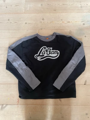 Svart och grå sweatshirt La Fam - Cool sweatshirt i svart med grå kontrastärmar och axelparti. Trycket 'La Fam' i vitt framtill ger en streetwear-vibe. Tröjan har rund halsringning och är tillverkad i mjukt bomullsmaterial. Perfekt för dig som gillar avslappnad stil.